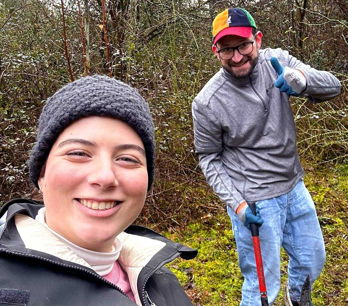 Two volunteers in front of some shrubs hold gardening tools and smile at the camera. 
